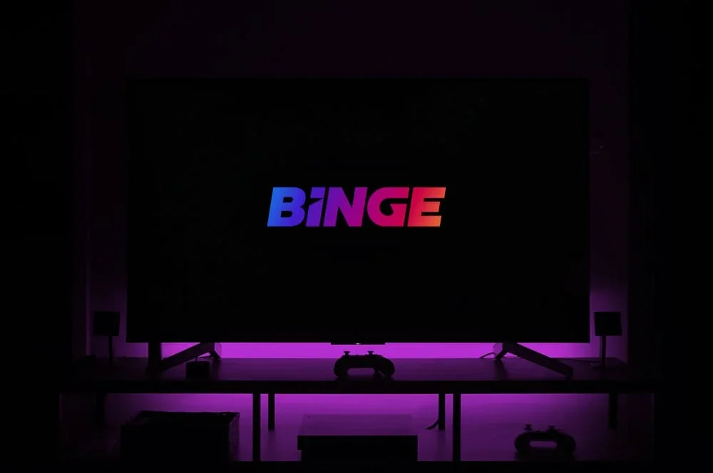 BINGE logo in gradient blue-to-red text displayed on a dark screen, with vibrant purple lighting below.
